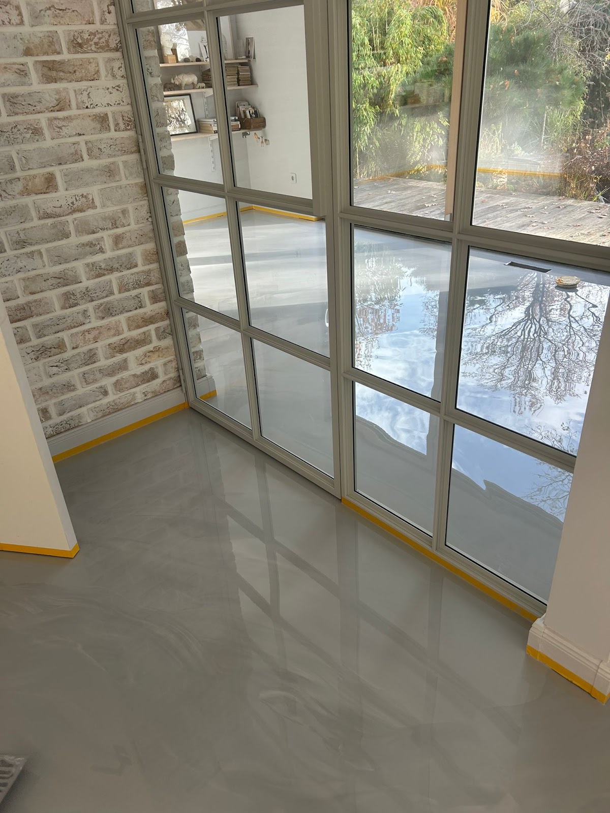 Moderner, glänzender Epoxidharzboden in grauer Marmoroptik mit bodentiefen Fenstern und einer Wand in Backstein-Optik.