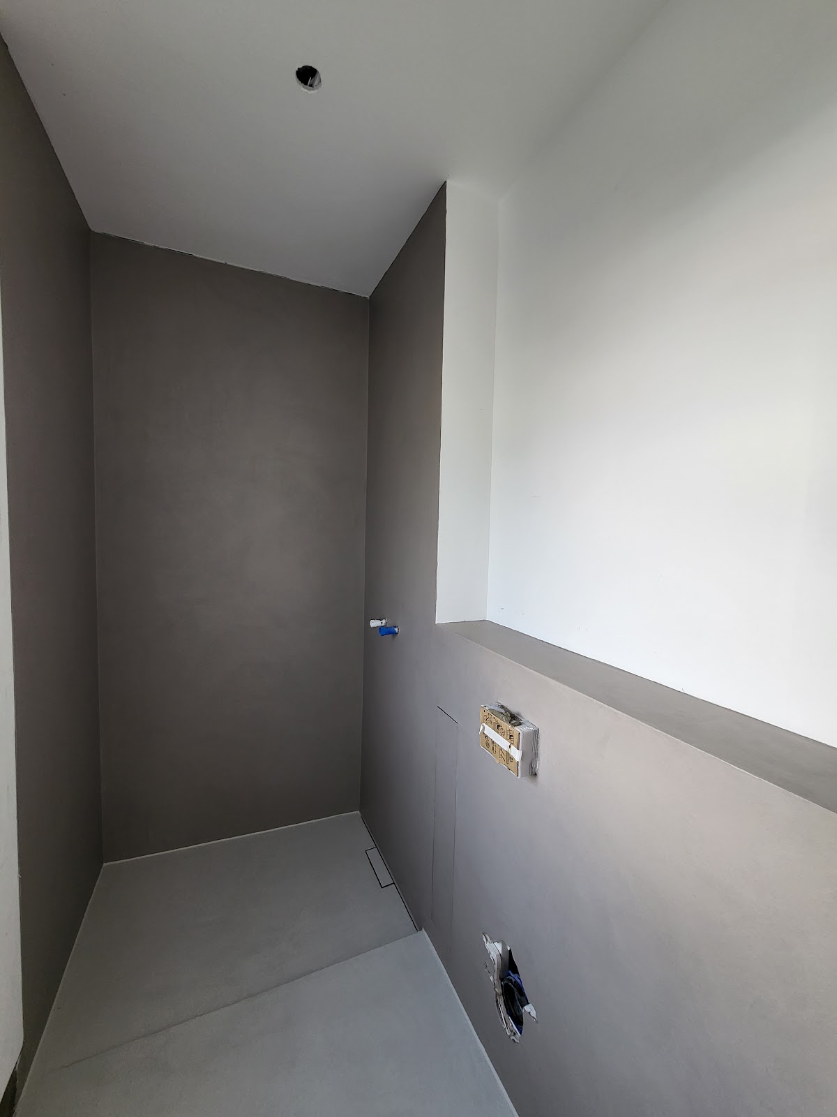 Teilweise fertiggestellte Dusche mit grauen gespachtelten Oberflächen und weißen Wandbereichen.