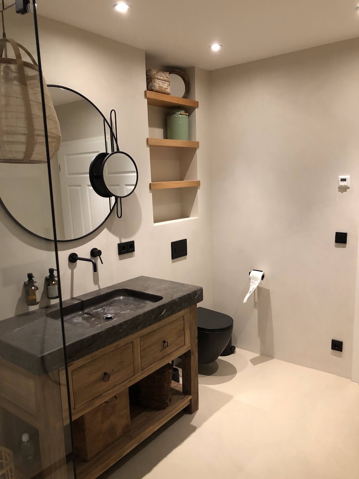 Modernes Badezimmer mit fugenlosen hellen Wänden und Böden, dunkler Waschtischplatte und schwarzen Armaturen.