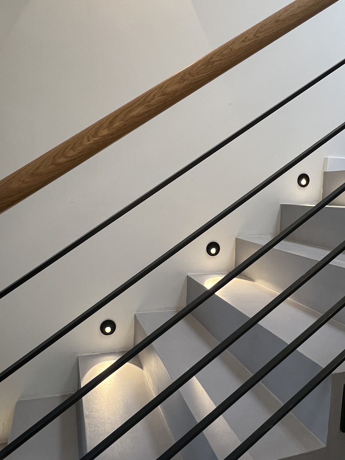 Moderne Treppe mit grauen Stufen, schwarzem Geländer, Holzhandlauf und Wandleuchten.
